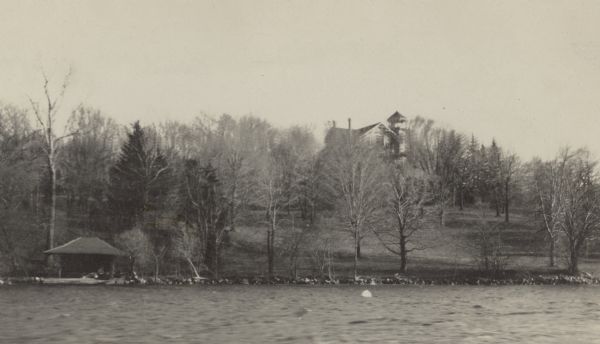 The main house at Black Point, with its distinctive tower, is seen through the leafless trees from Geneva Lake. The bath house sits at the water's edge. There is a buoy in the water near the shore.