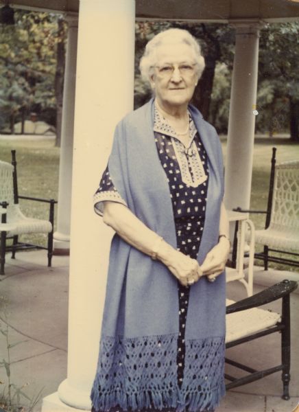 A handwritten note describes this portrait as: "Aunt Elsa in her summer house "The Lindens" when she was eighty years old." She is wearing a print dress and a knit, lightweight shawl and stands in front of an open columned gazebo with outdoor furniture.