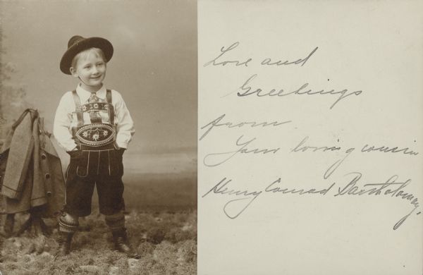Standing full-length portrait of Henry Bartholomay forms the left half of a postcard. The message reads: "Love and Greetings from Your loving cousin Henry Conrad Bartholomay." Henry is wearing a Bavarian style costume with lederhosen, shirt and tie, and hat. His jacket is hung on a prop on the left. There is artificial grass on the floor and a painted backdrop that includes an evergreen tree.