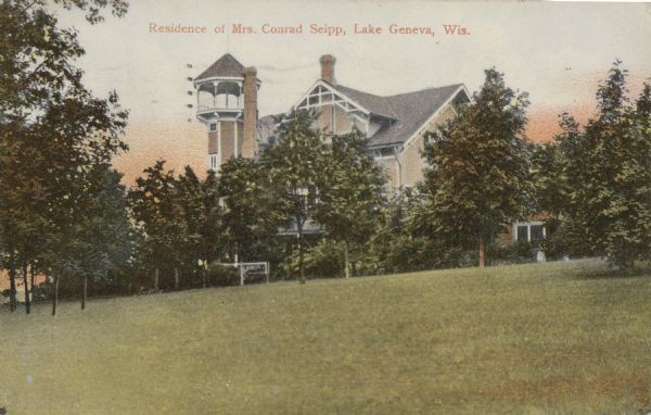 Hand-colored postcard bearing the inscription "Residence of Mrs. Conrad Seipp, Lake Geneva, Wis." The upper portion of the south side of the main house at Black Point, including the tower, is seen above the trees in the yard. Two tall chimneys and decorative stick work in the south gable are visible.
