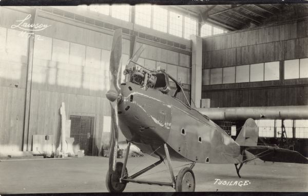 Photographic postcard of a three-quarter view from the front of the fuselage of a Lawson Military Tractor 2 (MT2) in a hangar.