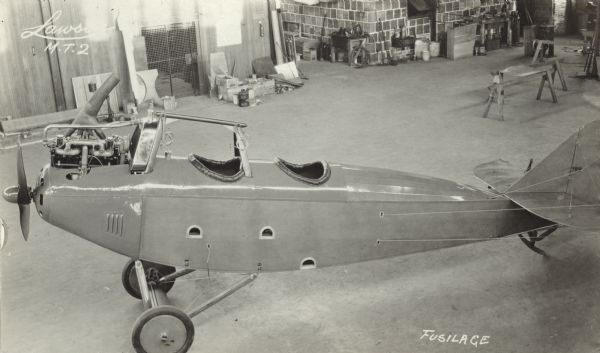 Left side of the fuselage of a Lawson Military Tractor 2 (MT2) in a hangar. In the background are tools, parts and work benches.