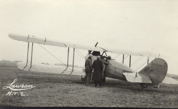 REar view from left side of a Lawson Military Tractor 2 (MT2) in a field. Three men are standing near the airplane, with one of the men leaning against the cockpit talking to the pilot.