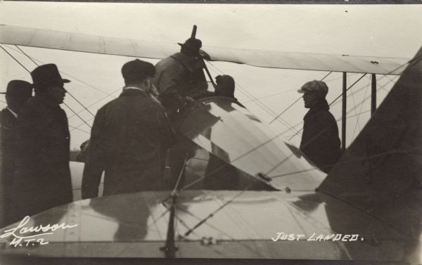View towards the cockpit of a Lawson Military Tractor 2 (MT2), apparently just after a successful flight. Five men are around the plane, and one man is standing up near the cockpit near the pilot. Location is unknown but is probably Lawson Aviation Field in Green Bay.