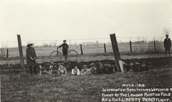 Fourteen children standing or lying down behind a barbed wire fence in a field, facing the camera. The postcard caption says they are watching an "MT2 for a Liberty Bond flight." Liberty Bonds were issued by the Aero Club of America around this time, to encourage development of new aircraft. Taken at the Lawson Aviation Field in Green Bay.