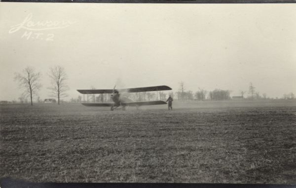 Lawson Military Tractor 2 (MT2) with the propeller engaged. A man is walking near the airplane's left wing, and the propeller and man are blurred. Location is unknown but is probably Lawson Aviation Field in Green Bay. Buildings are in the far background on the left and right.