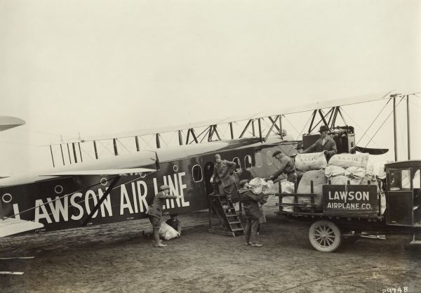Slightly elevated view of men loading bags marked "U.S. Mail" from a truck into a Lawson Airliner No. 2. Alfred Lawson in his flight gear is standing on a step ladder near the open hatch on the right side of the airplane, and another man is looking out from the hatch behind Lawson.<p>The caption attached to the photograph reads:
"THE UNITED STATES MAIL VIA THE MODERN AIRLINER
This is no doubt the greatest picture ever taken in the history of the Airplane. It shows how commercialism is now being introduced by Airplane in really and truly up-to-date fashion.
This picture shows the Lawson Airliner No. 2 which was designed by Alfred W. Lawson, the inventor of the first airliner, for the carrying of the United States mail for the Post-Office Department.
This Airliner is built to carry more than 6000 lbs. of mail at a time, and can fly from New York to Chicago during the night without making a stop.
This picture will give the public a practical idea of the tremendous strides made by the United States Post-Office Department in carrying the mail thru the air during the past three years.
When the Post-Office Department first started three years ago their airplanes only carried 100 lbs. of mail at a time, while this new modern leviathan of the air carries more than 6000 lbs. of mail at a time, or will carry an equal amount of mail in one load that it required sixty small airplanes to carry in the beginning of the Post-Office service.
This ship is powered with three Liberty Motors aggregating 1200 H.P. and has a wing spread of 120 feet. The body of this ship is 65 feet long and contains the main cabin in which a 7 foot high man can walk up and down with the same comfort and ease as he does in a railroad car. There is a mail chute in the bottom of this airliner thru which the mail is put into and taken out of the ship and this mail can be transferred from this ship to a smaller ship while it is flying thru the air and without the necessity of making a landing for the taking on or the delivering of mail to the different cities enroute. The cabin is sufficiently spacious to permit the mail clerks to sort the mail while the airliner is in flight."