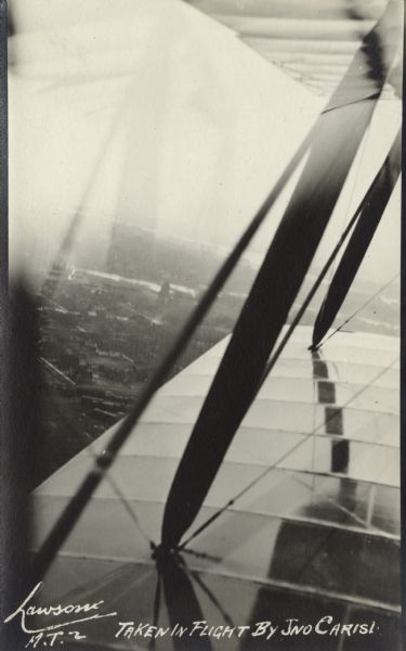 View over the wing of a Lawson Military Tractor 2 (MT2) in flight. Below are houses and what may be a river or lake. Location is unknown, but is probably near Green Bay. The photograph was taken by Giovanni Emmanuele Angelo Leonardo Carisi, who emigrated to the United States from Italy and was known as John Carisi. He served on the flight crews for some of Lawson's airplanes.