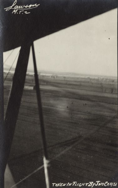 Photographic postcard of a view under the wing of a Lawson Military Tractor 2 (MT2) in flight. Below is a field and beyond are buildings and a body of water towards the horizon. Location is not identified but is probably near Green Bay. Photograph was taken by Giovanni Emmanuele Angelo Leonardo Carisi, who emigrated to the United States from Italy and was known as John Carisi. He served on the flight crews for some of Lawson's airplanes.