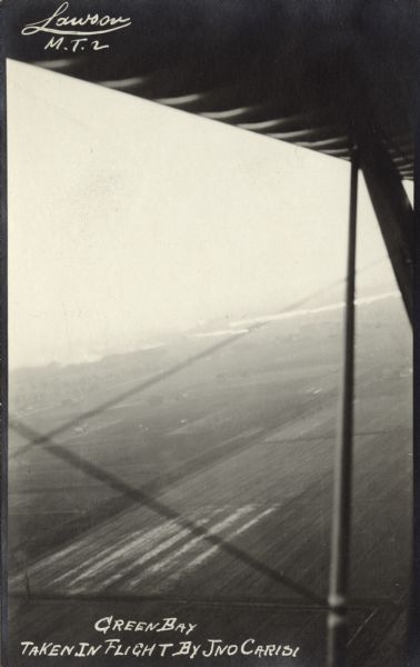 Photographic postcard showing of a view behind the wing of a Lawson Military Tractor 2 (MT2). Below are fields and in the distance are houses. The photograph was taken by Giovanni Emmanuele Angelo Leonardo Carisi, who emigrated to the United States from Italy and was known as John Carisi. He served on the flight crews for some of Lawson's airplanes.