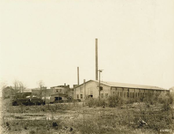 Exterior view from field across pond towards Alfred Lawson's aircraft plant, with buildings and smokestacks. Men are working with a pile of lumber on the left. 