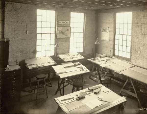 Elevated view of drafting tables for aircraft design, presumably inside the Lawson plant. There are several stools, desks and tables set up in the corner of a room with three windows. Papers, rulers, pencils, and other drafting tools are on the tables. A framed print of an airplane is on the wall, beneath a "No Smoking" sign.