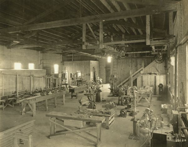 Interior of the machine shop at the Lawson plant. There are drill presses and a belt-driven machinery system attached to the open rafters of the ceiling. Several airplane parts are on tables and saw horses. In the back of the room near a window to an office is a sign for the "Adams Express Company." Near a washbasin under a window on the right is a box of Fairbanks Gold Dust Washing Powder.