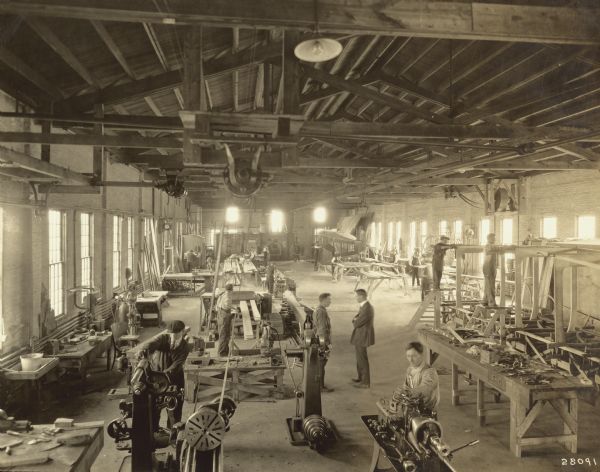 Interior view of the Lawson plant. At least 18 people are working in the large, open room. Belt-driven machinery is attached to the rafters of the open ceiling. In the rear of the shop is a partially assembled fuselage.
