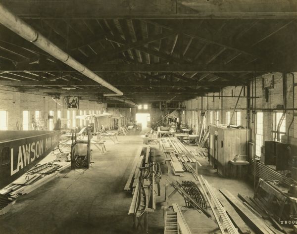 Elevated view of the interior of the Lawson plant, a large open room with exposed ceiling and windows lining both walls. Part of the fuselage for a Lawson Air Liner is on the left against the wall. Airplane parts are laid out across sawhorses, and on the ground. A small, partitioned room on the right has a "No Smoking" sign, and two doors. 