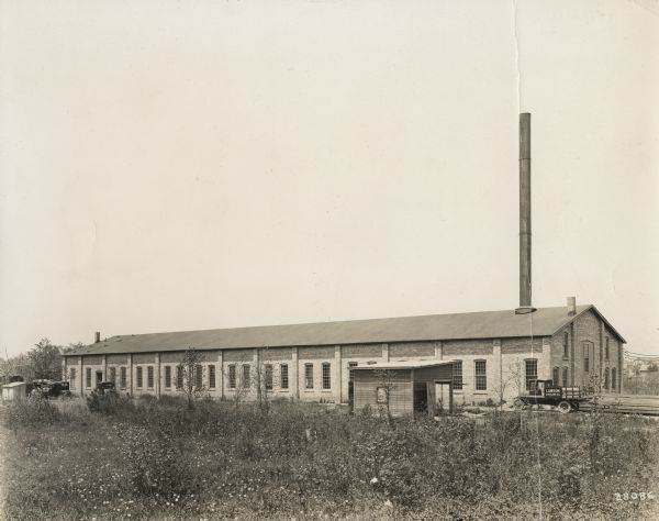 Exterior view from field of a long brick Lawson plant building with a smokestack. Several automobiles are parked near the plant on the left, and on the right is a truck with "Lawson Airplane Co." painted on the side. There is a small outbuilding in the center foreground. Houses are in the far background.