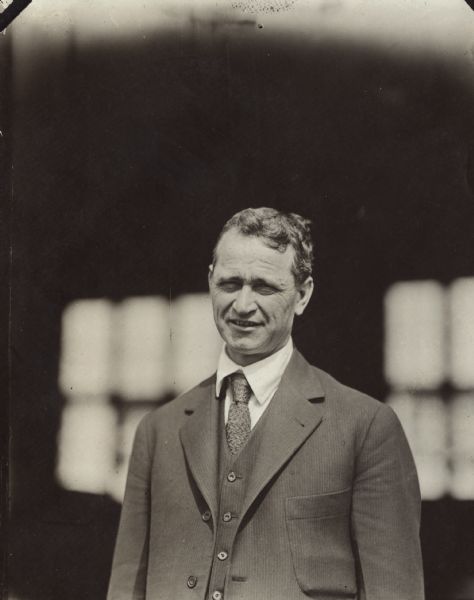Waist-up portrait of Alfred Lawson standing, wearing a suit. An unidentified building is behind him. Caption on the back of the photograph reads: "Return to Mr. A.W. Lawson, Milwaukee Athletic Club, Milwaukee, Wis. Alfred W. Lawson, Designer and Navigator of the Lawson Air Liner."
