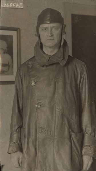 Three-quarter length portrait of Alfred Lawson standing indoors. He is wearing a long leather coat and cap. 