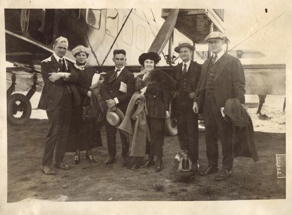 Group portrait of Alfred Lawson, two unidentified women, and three unidentified men. They are standing outdoors in front of a Lawson Air Liner. A leather satchel is on the ground in front of one man.