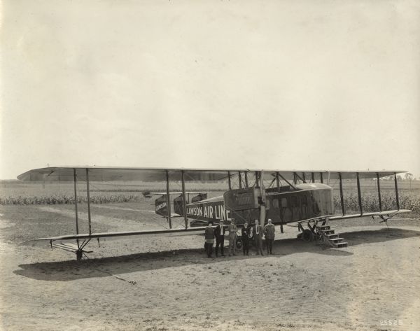 Elevated view of a group of five men and one woman standing in front of a Lawson Air Liner parked in a field. 
