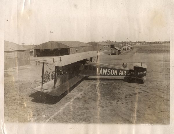 Elevated view of several men, most in military uniforms, inspecting a Lawson Air Liner outside a hangar at an airfield. The hangar door says 51st AERO SQDN. Although the location is unidentified, the timing of the plane and the locations of the 51st Aero Squadron indicate that this is probably 1918 and is either at Ellington Field in Texas or at Henry J. Damm Field in New York.