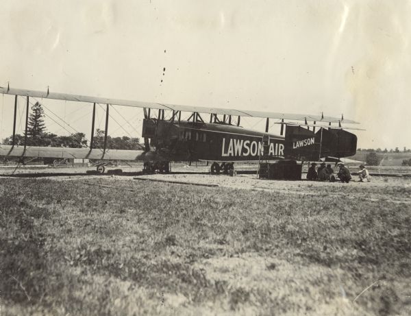 View across field towards a Lawson Air Liner sitting in a field. Five men are crouched or seated underneath its tail assembly. A ladder is propped against the airplane.
