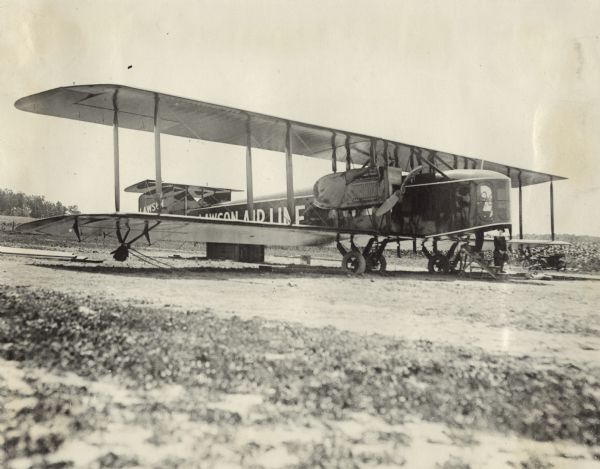 Three-quarter view from front right of a Lawson Air Liner. A woman is standing on the ground near the cockpit. A man is standing on wooden steps that lead into the cockpit.
