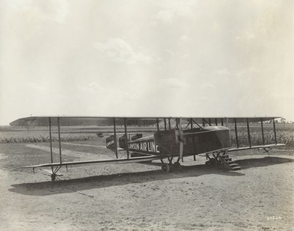 Slightly elevated three-quarter view from front right of a Lawson Air Liner parked in a field. A small set of wooden steps are positioned in front of the left wing, near the cockpit.