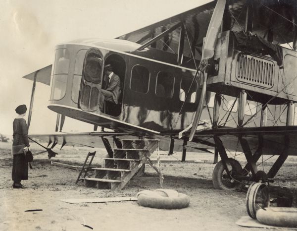 Close-up three-quarter view from front left of the cockpit of a Lawson Air Liner. A small set of wooden stairs is positioned near the cockpit. A man is seated in the cockpit with his head turned away from the camera. A woman is standing in front of the cockpit on the left.