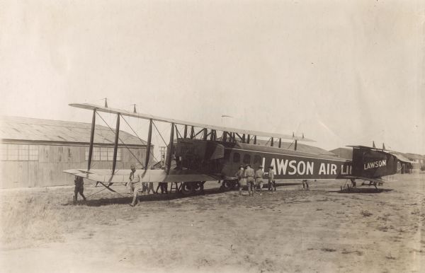 Three-quarter view from the right rear of a Lawson Air Liner. Several men in uniforms are standing near the airplane. Behind is a hangar with 51st AERO SQDN written on the side.
