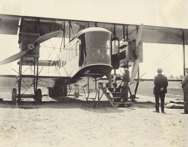 Close-up of the cockpit of a Lawson Air Liner. Two men are standing on the right in front of the airplane, and a woman is standing on the lowest step of a set of wooden stairs that lead up into the cockpit. A man is standing at the top of the stairs looking into the cockpit.