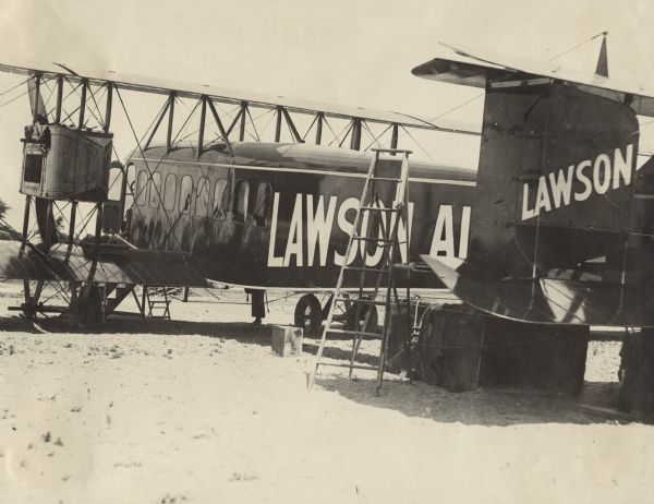 Three-quarter view from left rear of the Lawson Air Liner sitting in a field. A ladder is standing near the tail of the plane. Wooden steps and a chair are underneath the cockpit.