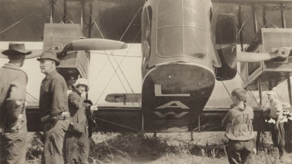 Two men, a woman, and two boys are standing in the foreground in front of a Lawson Air Liner. Part of the word "LAWSON" is painted on the underside of the airplane.