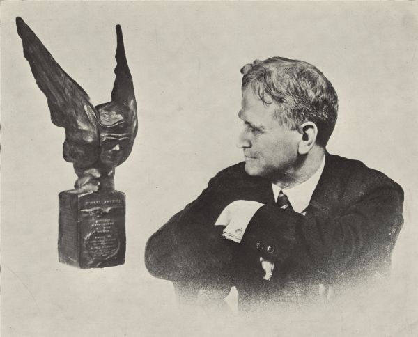 Vignetted waist-up portrait of Alfred Lawson, looking at the "Winged America" trophy he received from the Aerial League of America. The trophy depicts a pilot's head cap a goggles, and wings framing the face.