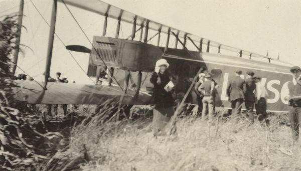 Several people standing  around a Lawson Air Liner, which is in profile. Tall grasses are in the foreground.