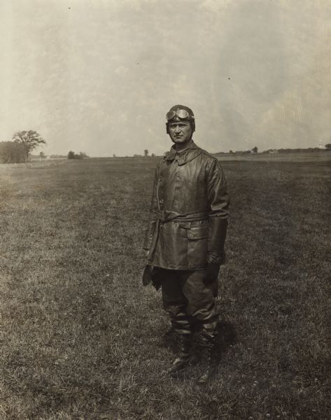 Full-length portrait of Alfred Lawson standing in a field in leather flight gear. This is around the time the Lawson Aircraft Company built the Military Tractor planes, in Green Bay.