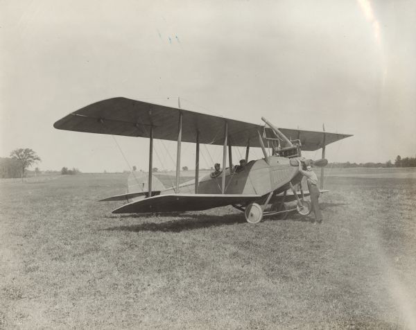 Alfred Lawson is seated in the rear compartment of a Lawson Tractor Biplane, which is parked in a field. A man in a suit is seated in the front compartment. A third man poses with his hands on the propeller.