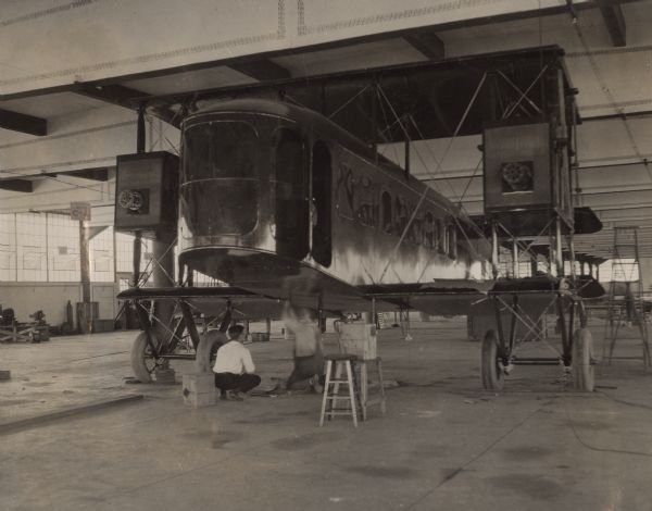 Exterior plates being attached to a Lawson Air Liner. Two men are crouching underneath the cockpit.