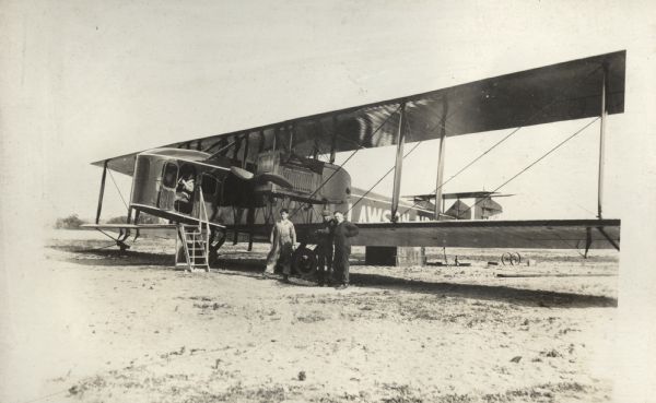 Three men in work uniforms stand next to a Lawson Air Liner, which is parked in a field. A fourth man is seated in the cockpit.