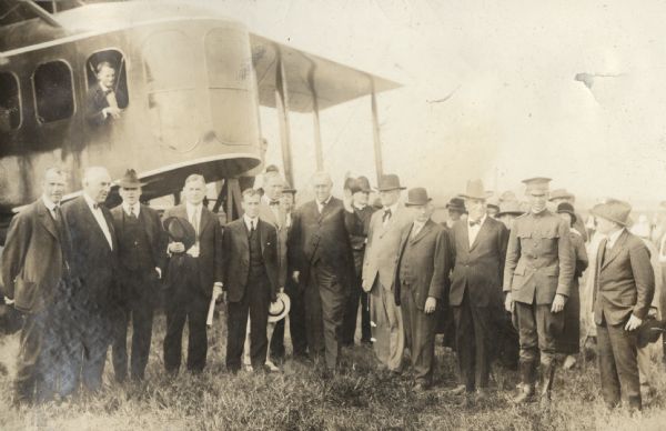 A group of well-dressed men and women stand in front of a Lawson Air Liner, which is parked in a field. One man is wearing a military uniform. Another man is leaning out of a window on the plane.