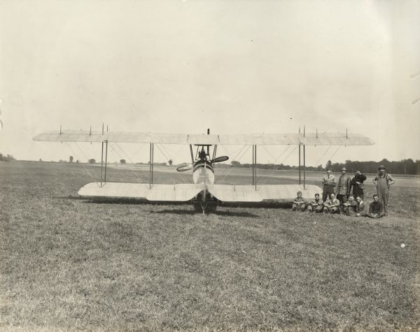 Rear view of a Lawson Tractor Biplane, which is parked in a field. The flight crew stands and sits to the right of the plane. Alfred Lawson is in the center, in his leather flight gear. Giovanni "John" Carisi stands to his right.