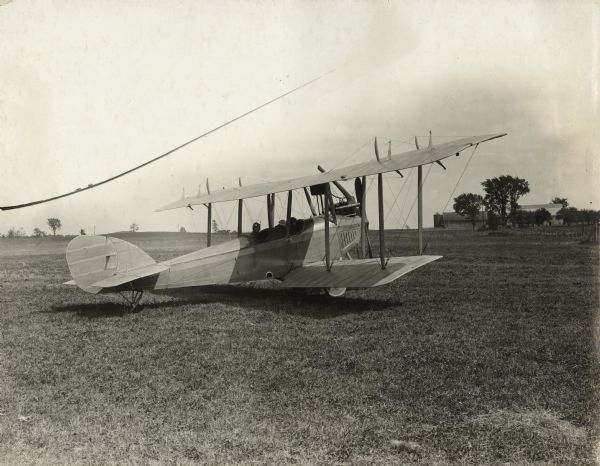 Alfred Lawson is seated in the rear compartment of a Lawson Tractor Biplane. The front compartment passenger is barely visible. A farm is in the distance.