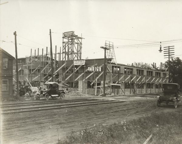 A building is under construction, and features a banner that reads: To Be Occupied By Lawson Aircraft Corp. A sign on part of the construction reads: Ludolf M. Hansen Co. Contractors. Two cars are parked in front of the building, and railroad tracks are also in the foreground.