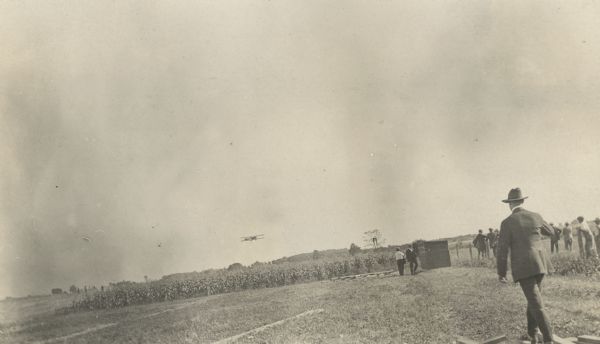 A Lawson Air Liner is shown in flight above a field. In the foreground, several people stand along a fence, watching the plane.