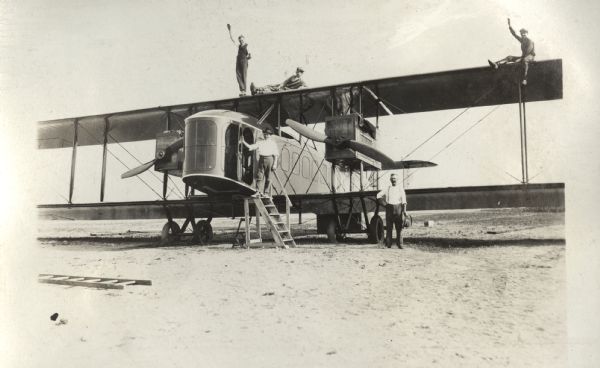 Five men posing with a Lawson Air Liner. Three men are standing on the upper wing of the plane. There is a man standing on a ladder at the cockpit.