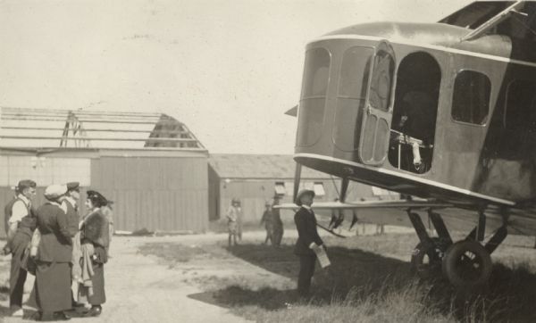 A Lawson Air Liner parked near a hangar. Men in uniforms are in the background, and suggest this could be a military base. Several people are standing nearby, some looking at the plane. One man is standing underneath the cockpit, looking up at it.