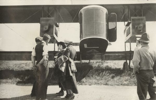 Six people are standing in the foreground, with a Lawson Air Liner behind them. One of the men has his back to the camera, but might be Alfred Lawson. The plane's propellers are in motion.