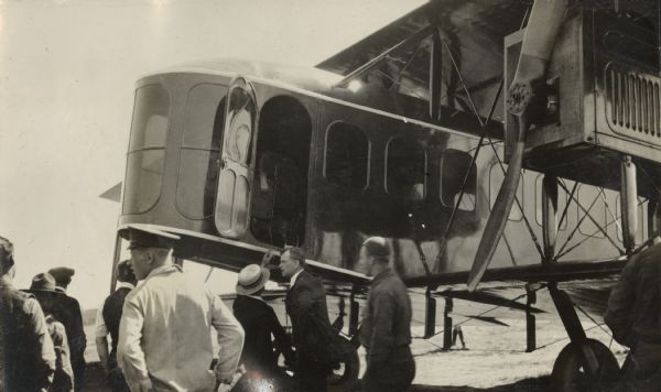 Several people are standing in the foreground, with a Lawson Air Liner behind them. Through the cockpit door, the pilot's seat and safety belt are visible.