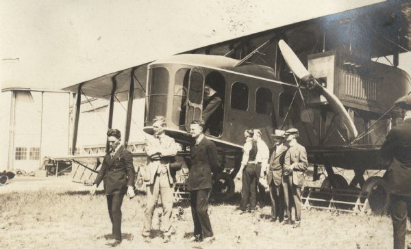 Alfred Lawson standing with his arms folded, between Giovanni "John" Carisi and another man. Behind them, other men are standing near a Lawson Air Liner.