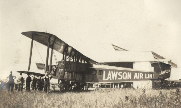 A Lawson Air Liner is parked in front of a building that might be a hangar. Several men are standing near the plane, which is visible from its left side.